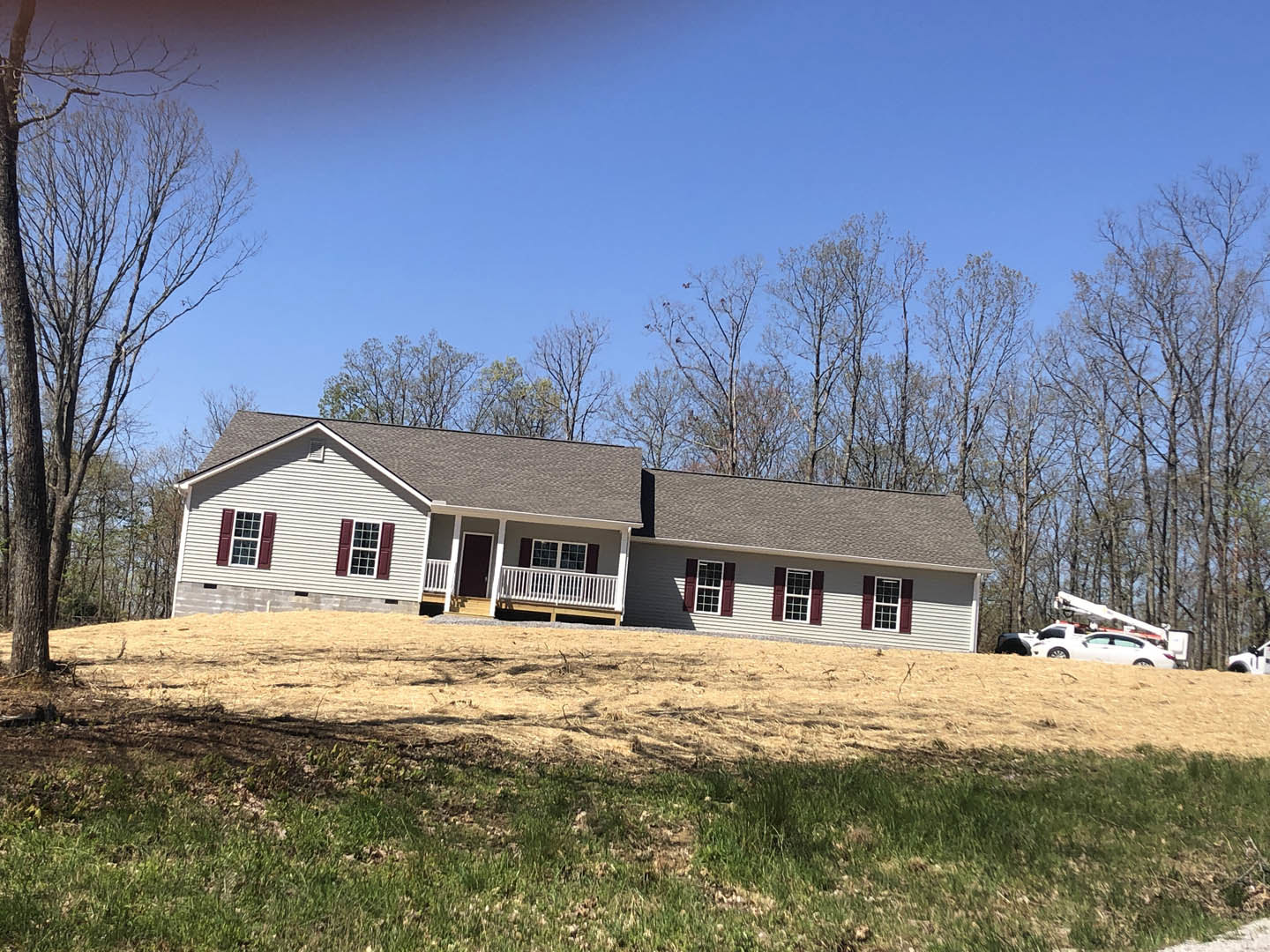 White-roofed house with covered porch, parked car on grassy hill, leafless tree and multiple square windows, surrounded by open lawn and trees in the background