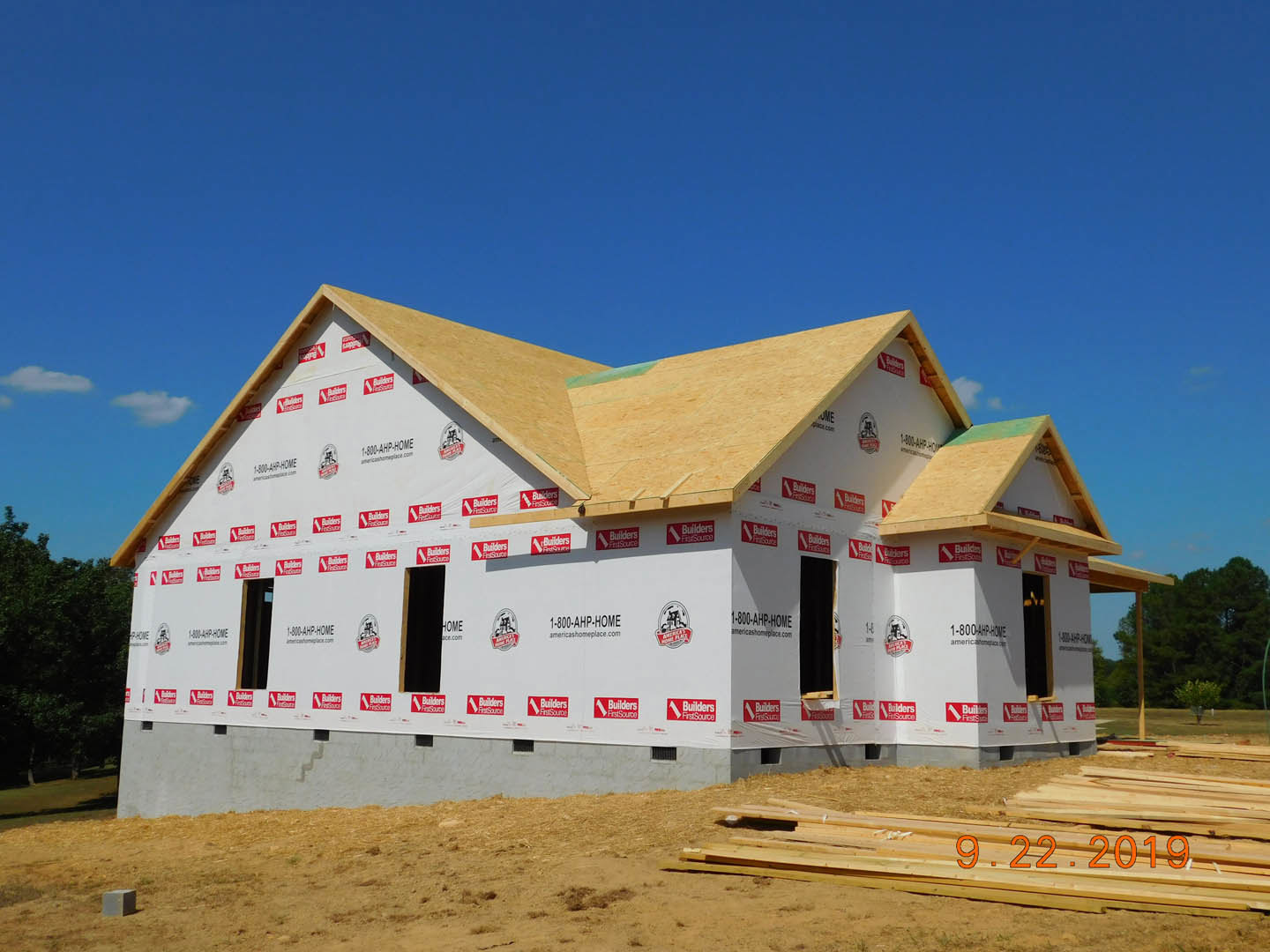 Framed house under construction with exposed wood planks, red and white branded wrap, blue sky overhead, pile of numbered lumber, white pole with red stripes, black rectangular