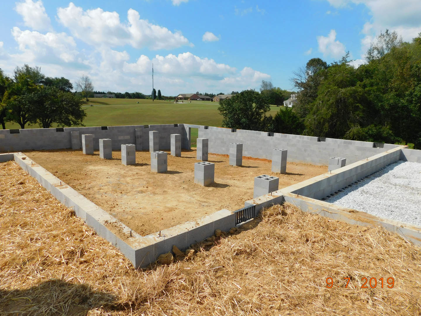 Concrete foundation with cement pillars set in dirt, surrounded by grass and trees under a blue sky with scattered clouds.