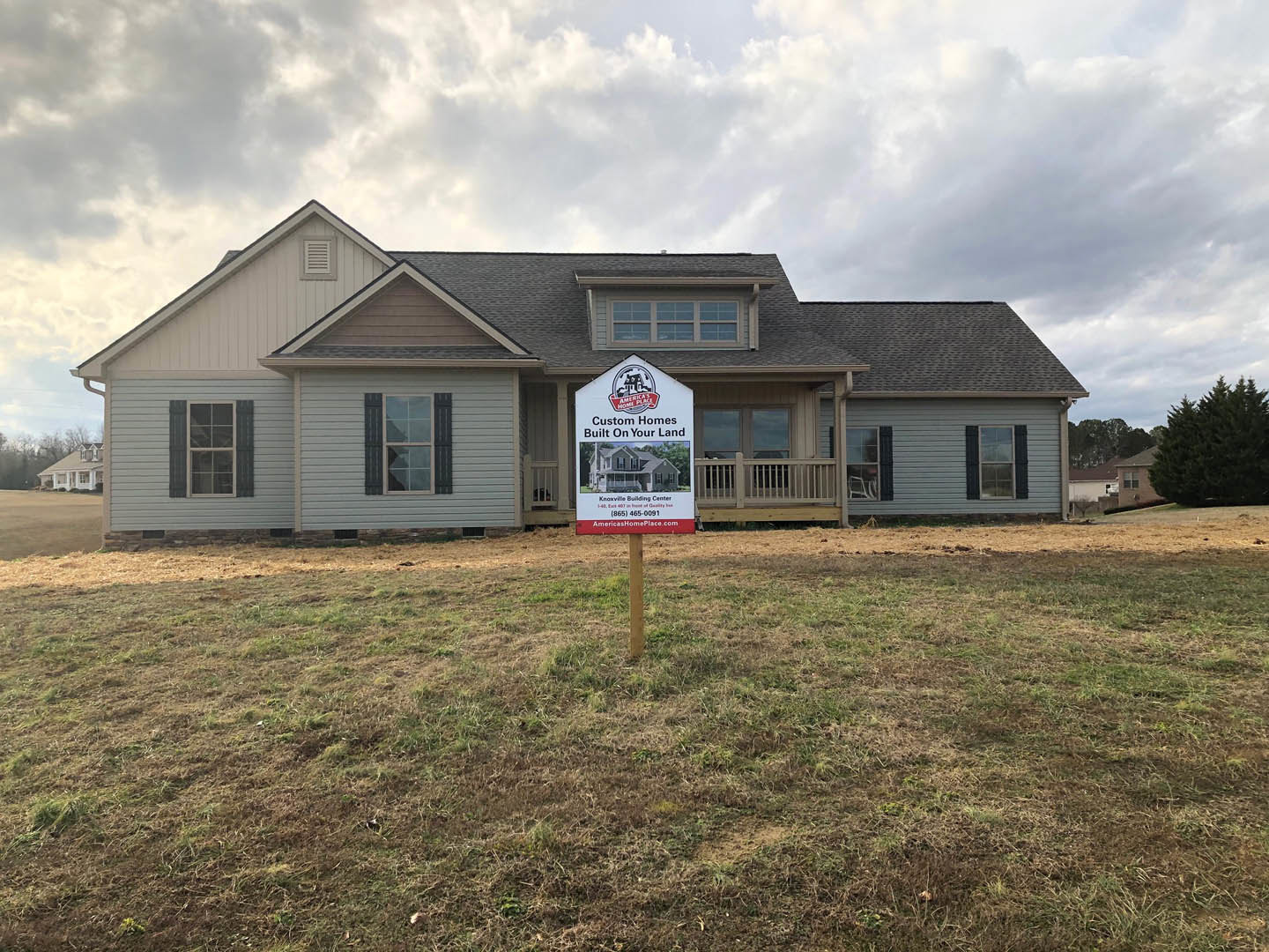 Two-story custom home with white siding and black-framed windows, manicured lawn, and a real estate sign in the front yard; mature tree and cloudy sky in the background