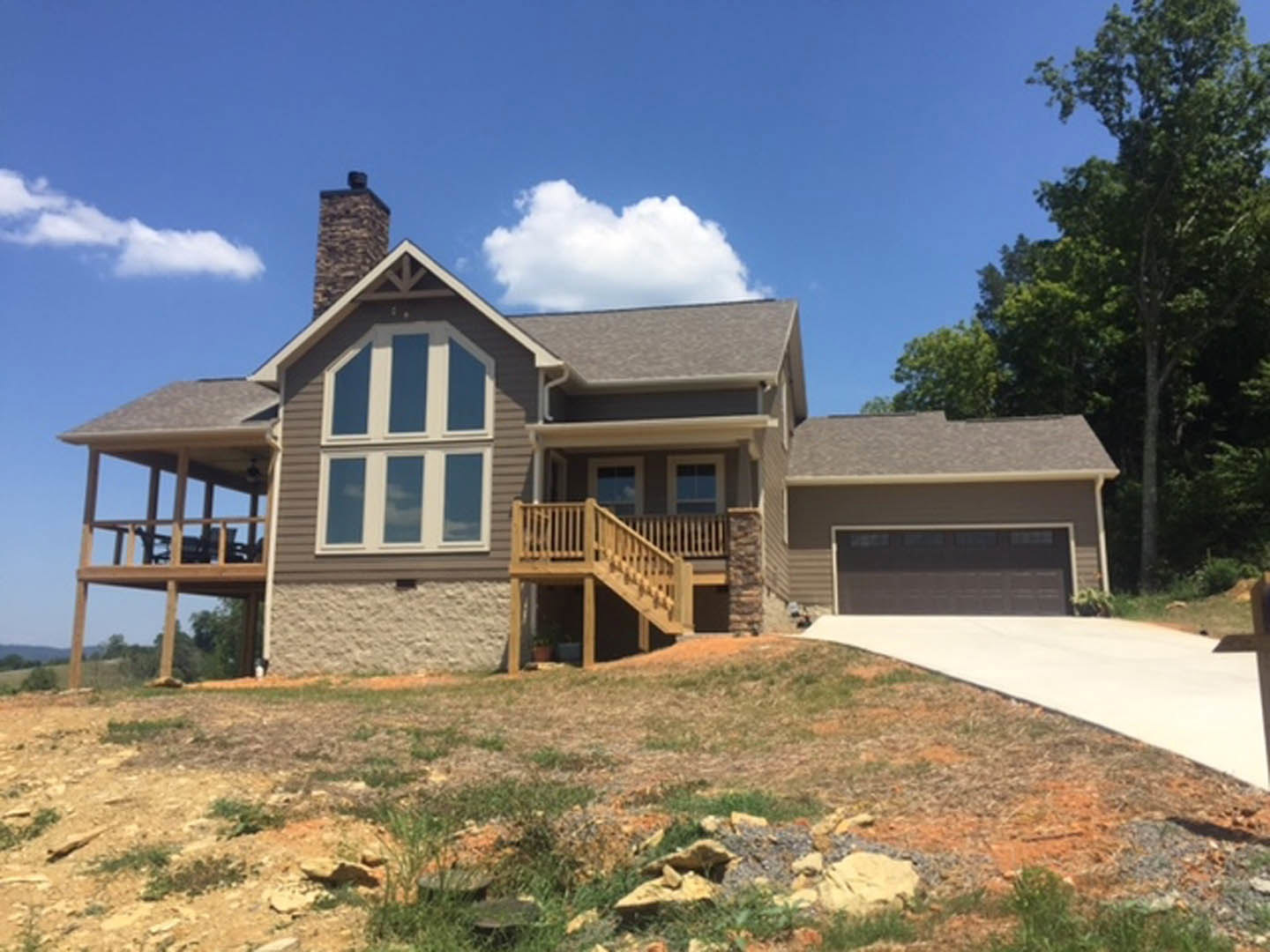 Two-car driveway leading to a modern home with light siding, wooden deck with railing, large windows, and landscaped front yard with mature trees under a partly cloudy sky