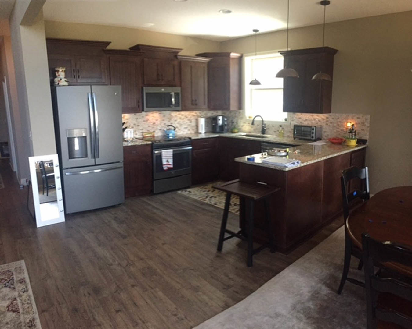 Kitchen with wood flooring, central dining table, white cabinetry, stainless steel refrigerator, oven with towel draped on handle, microwave on countertop, and patterned rug