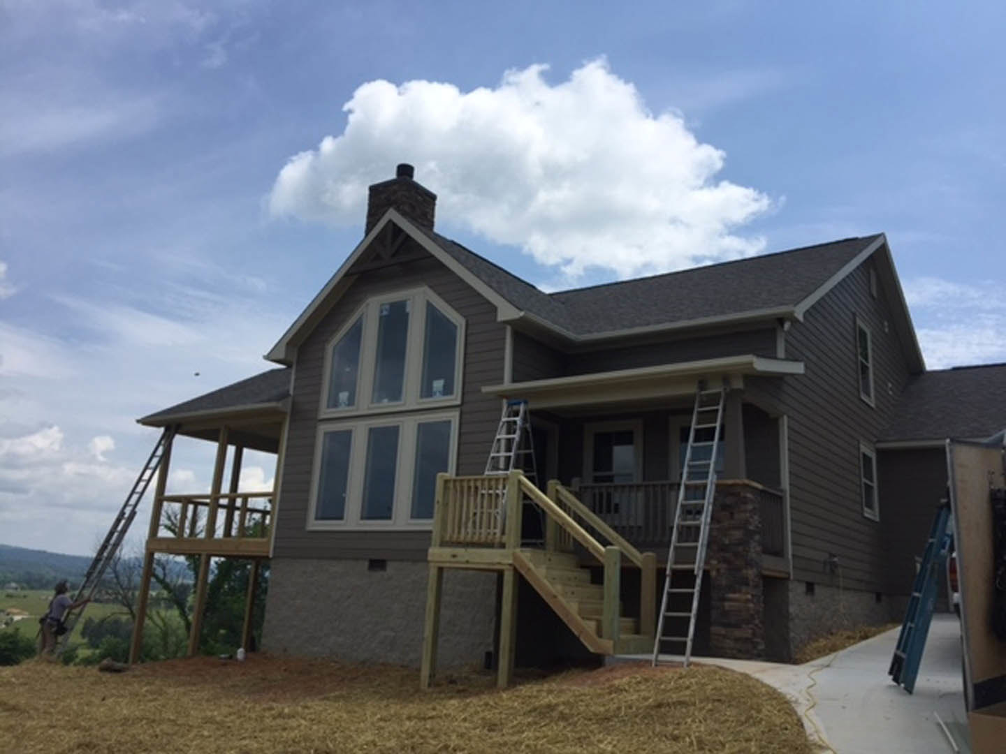 Two-story house with white siding, wooden exterior stairs, ladder leaning against side wall, grassy lawn in foreground, porch, windows, and chimney under partly cloudy sky