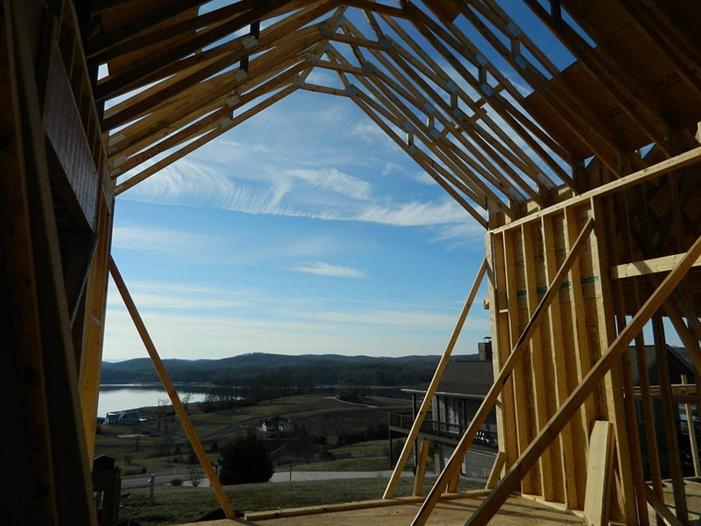 Wood-framed house under construction overlooking a lake and distant hills, exposed beams and roof structure visible, cloudy sky above