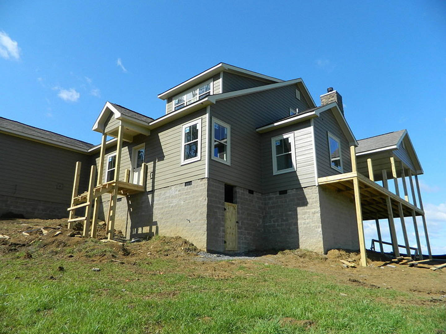 Partially built house with exposed wooden staircase, unfinished exterior walls, wooden door, and window displaying construction signage, surrounded by grass.