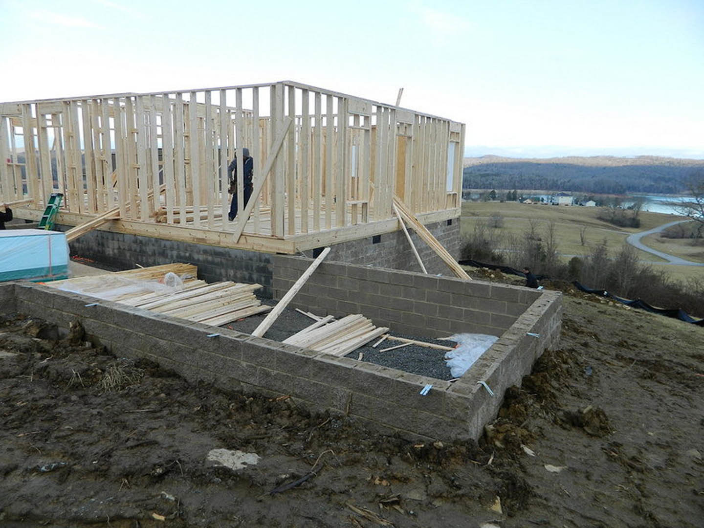 Wood-framed house under construction on a hillside, blue tarp draped over part of the structure, pile of lumber on dirt ground, cloudy sky in background