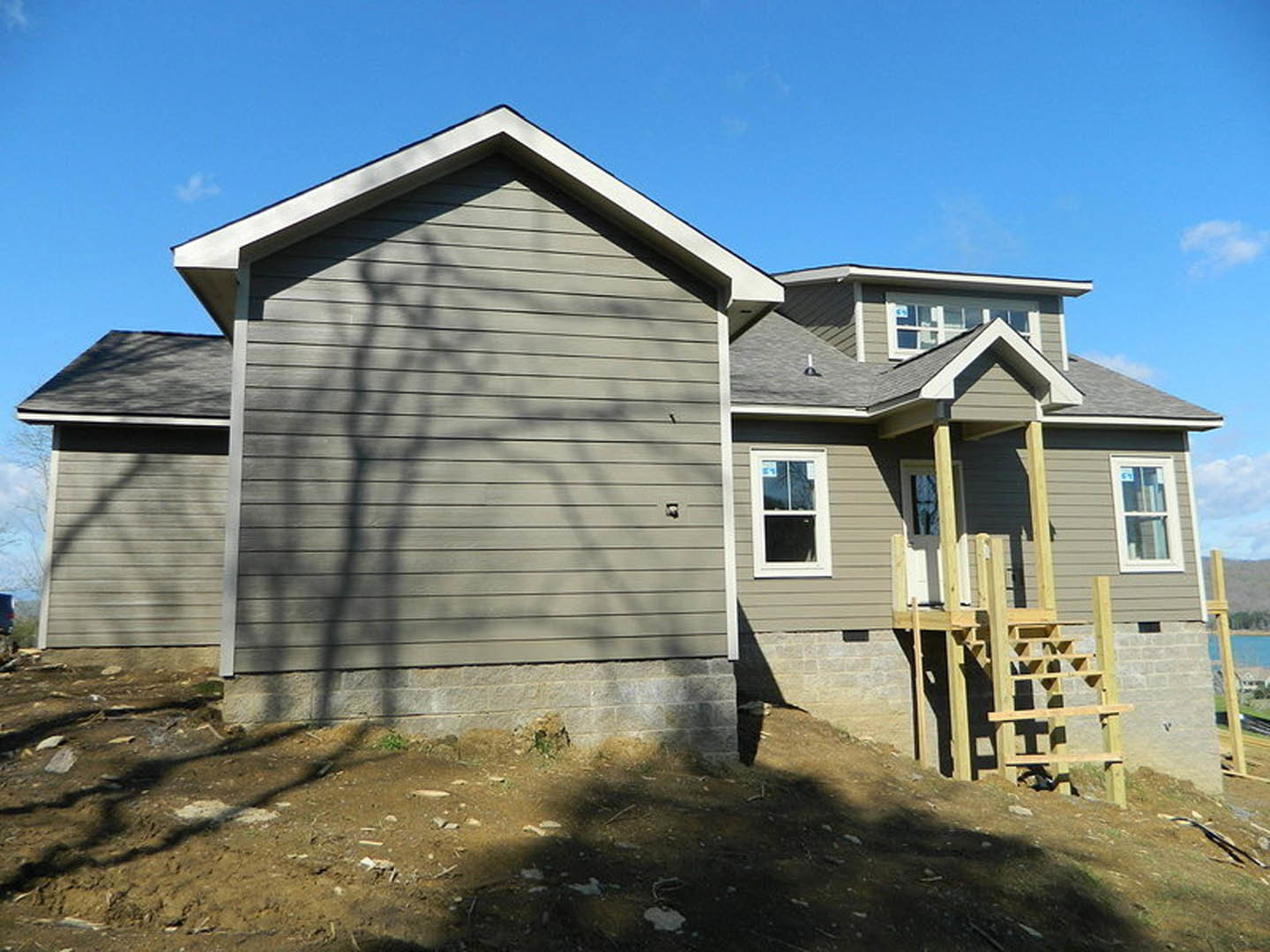 Two-story cottage-style home with light siding, large windows, covered front porch, wooden exterior staircase, and tree shadows cast across the facade