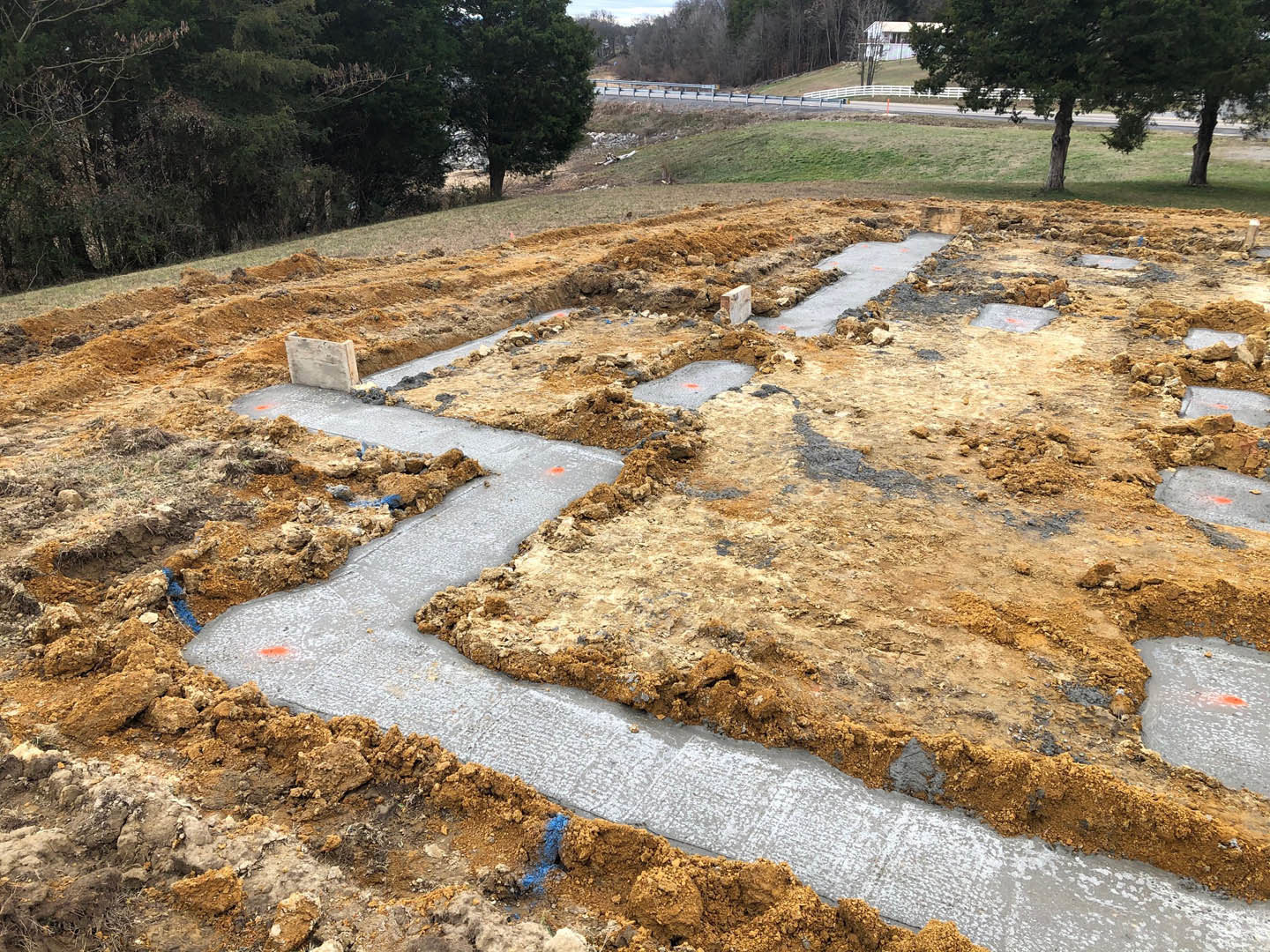 Dirt construction site with poured concrete footers, wooden board walkway, grassy field, and tree in background