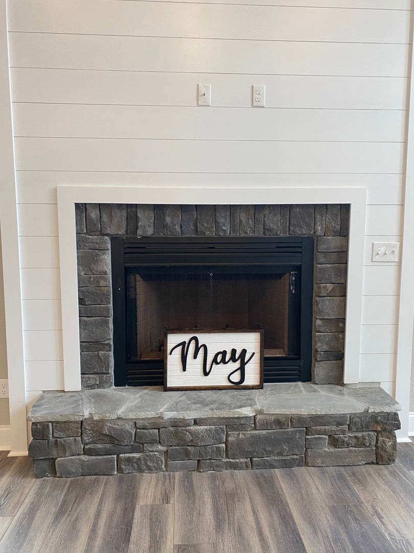 Stone fireplace with wood mantel, decorative sign displaying a name, hardwood floor, fire screen, and hearth in a living room.