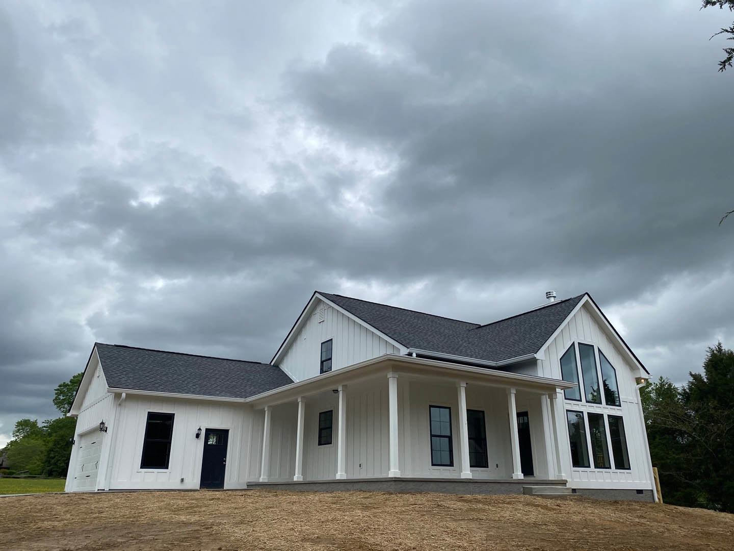 White two-story house with black-framed windows, white columns, and a cloudy grey sky; trees and grassy field in the background