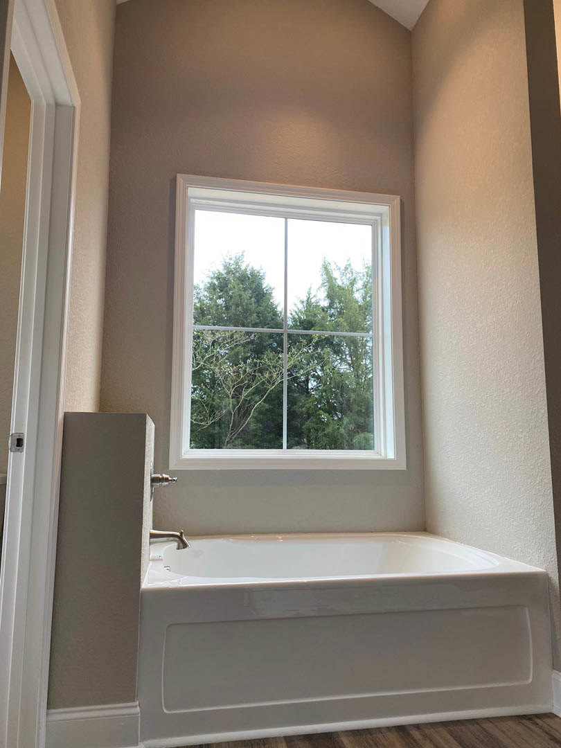 Freestanding white bathtub on wood floor beside large window with tree views, white walls, and minimal trim detailing