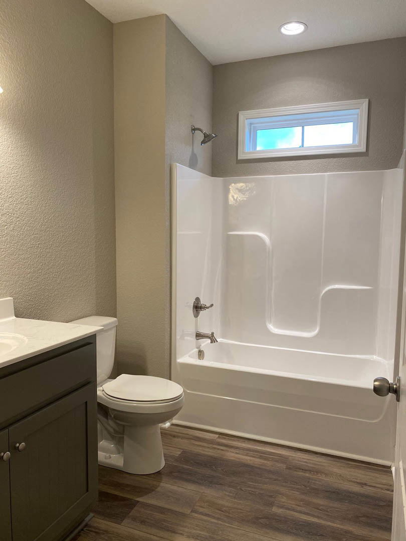 Modern bathroom featuring a white bathtub, closed toilet, wood floor, tile walls, large window with blue sky, and built-in cabinet