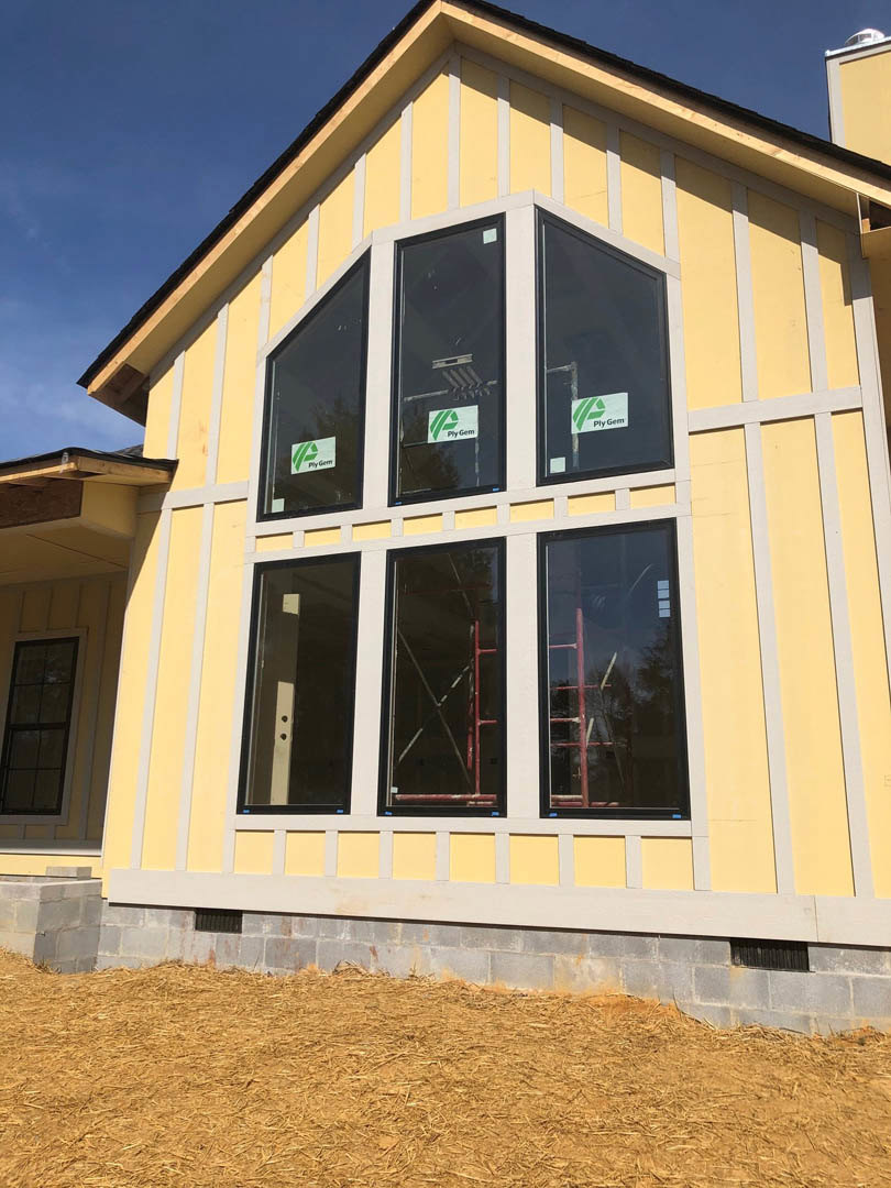 Yellow house under construction featuring multiple black-framed windows, exposed siding, ladder leaning against wall, and straw pile on ground