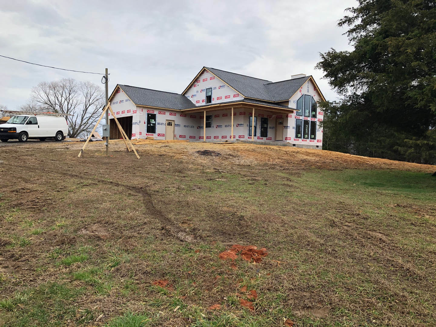 Partially built house with exposed framing, surrounded by a large grassy field, white van with roof ladder parked nearby, leafy tree, pile of dirt, construction signs attached to