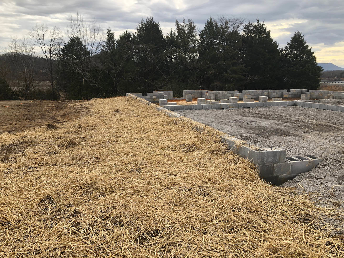 Concrete foundation surrounded by grass, hay pile, and stacks of grey blocks; leafless tree and group of trees under cloudy sky in background