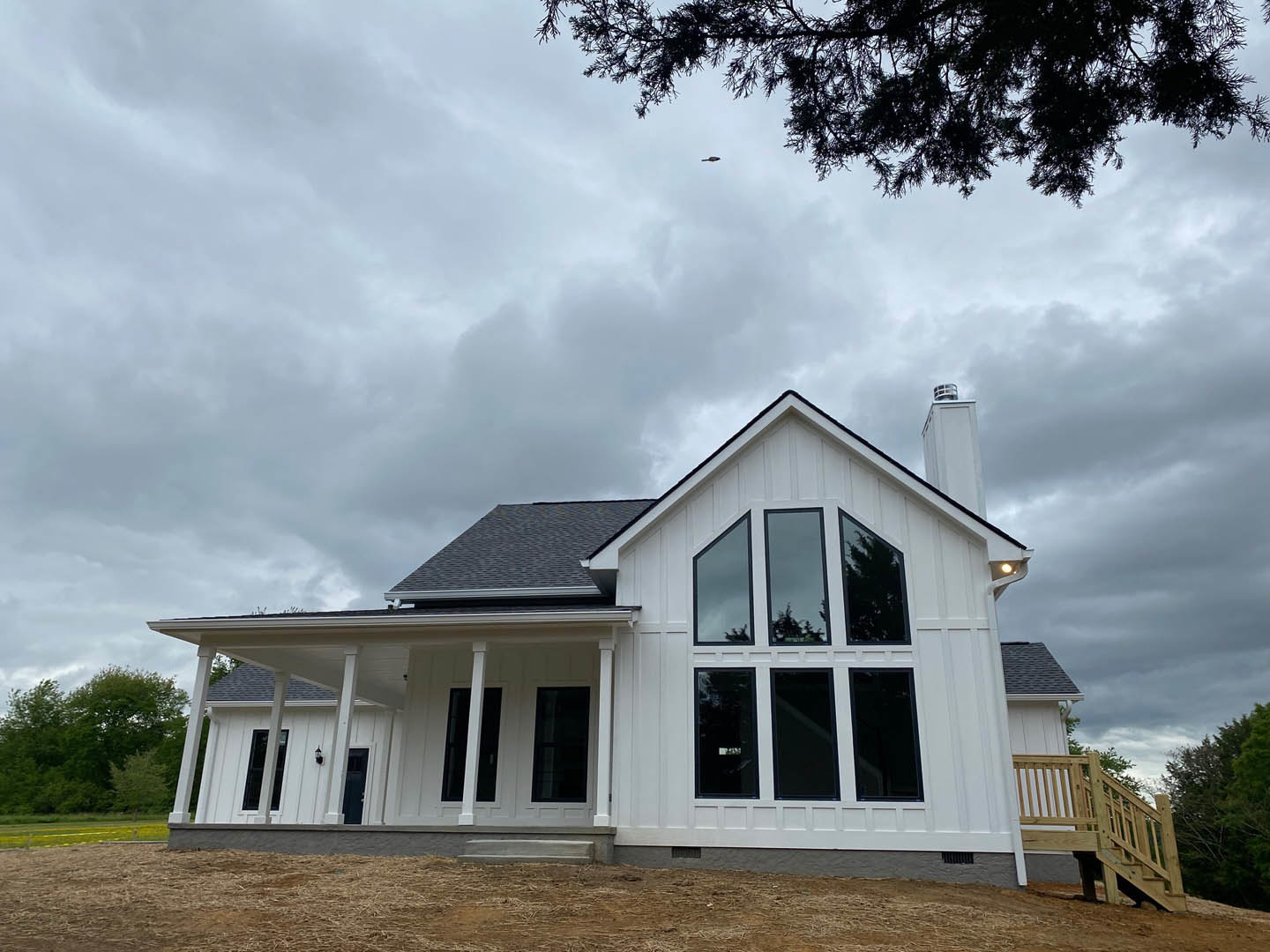 White siding house with large brick chimney, covered front porch, wooden steps, and mature trees in the background under overcast sky