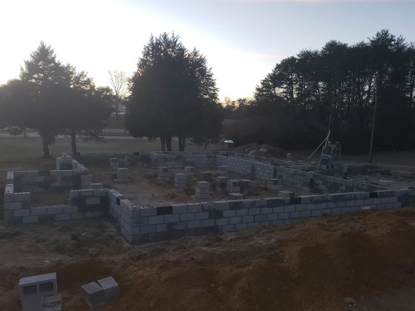 Concrete foundation surrounded by stacks of grey bricks, pile of dirt, and leafy tree on a residential building site