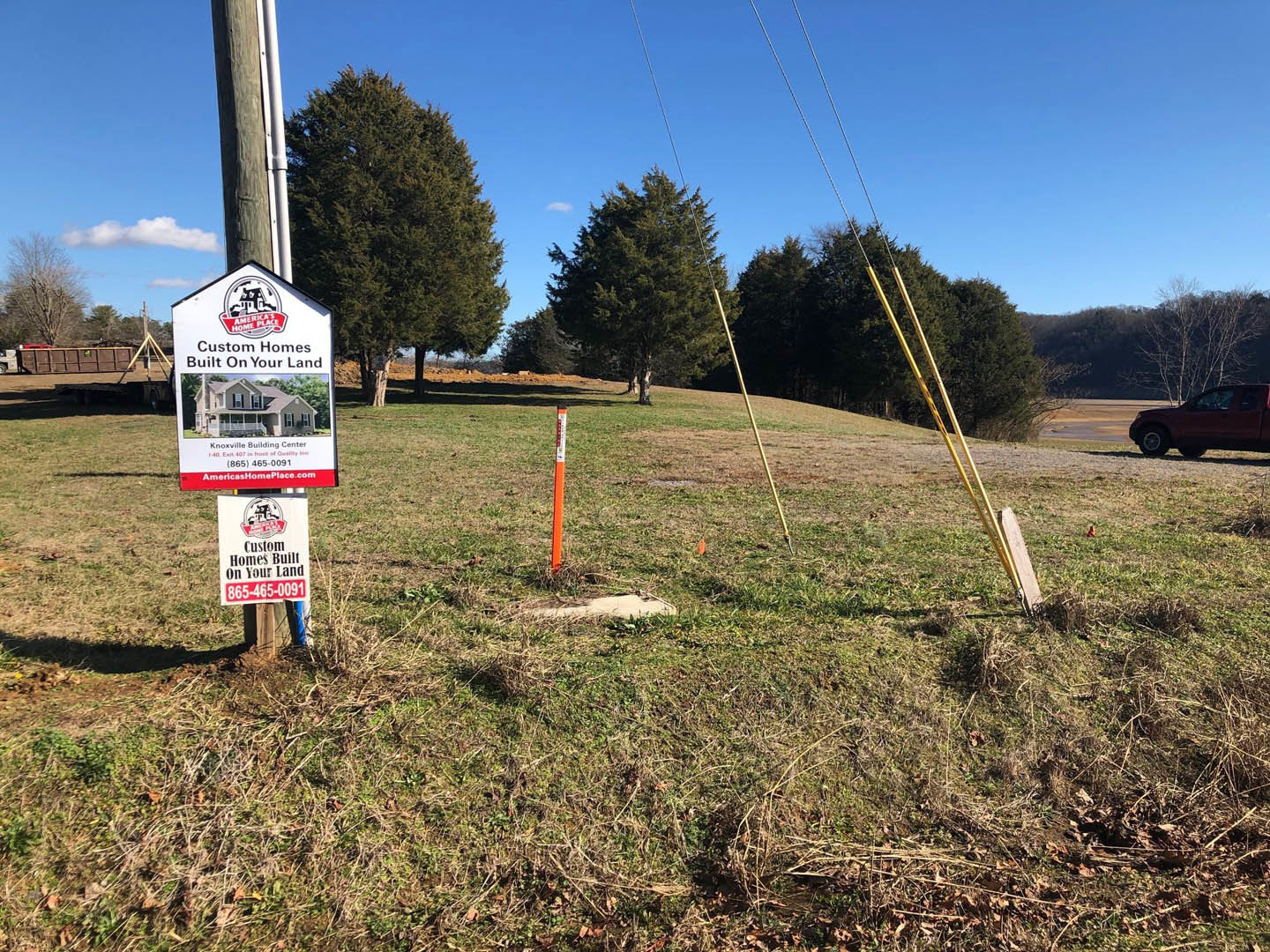 Wooden sign with house illustration mounted on a pole in grassy field, large leafy tree nearby, red truck parked in background under blue sky.