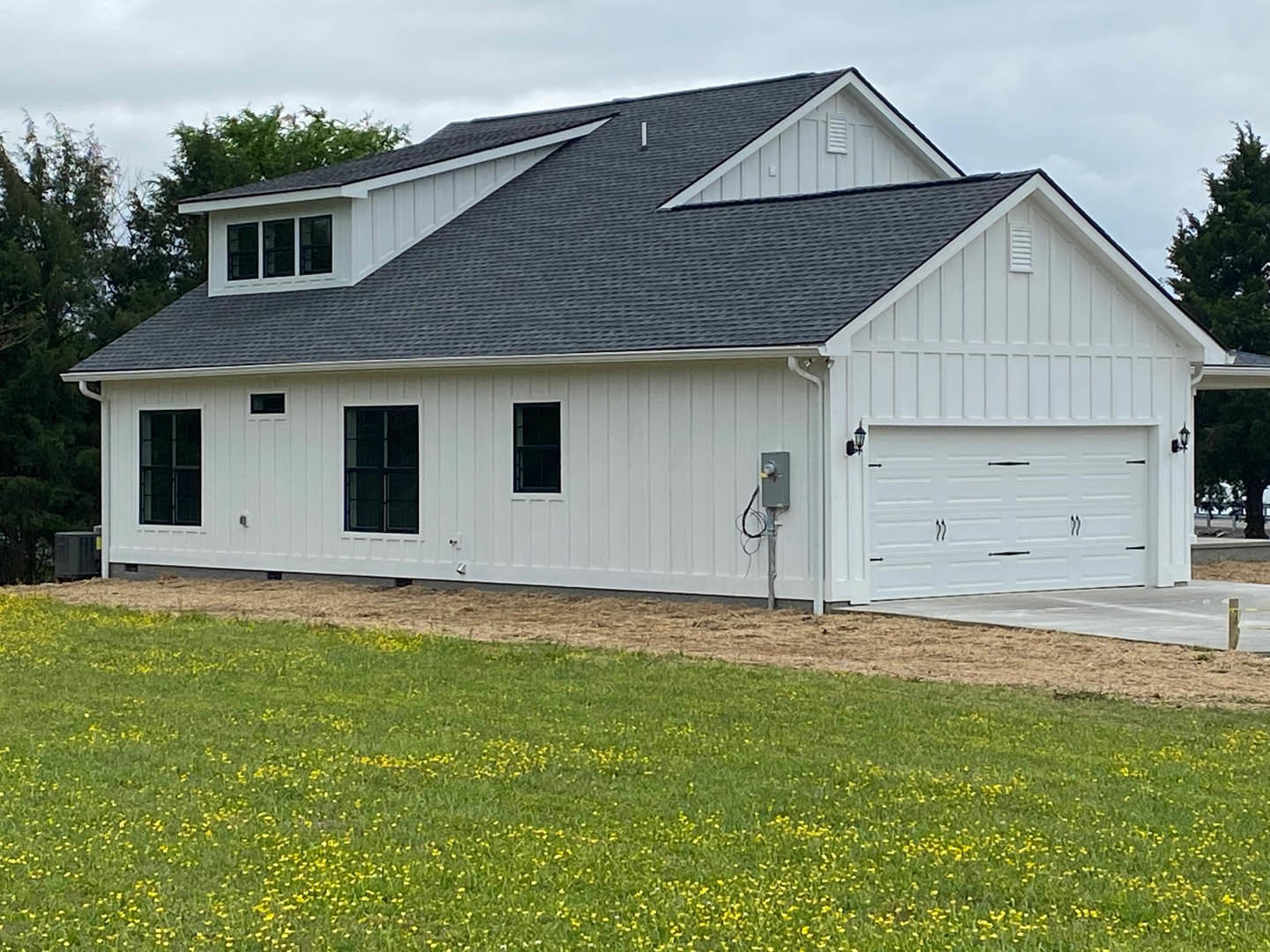 White farmhouse with black roof, white-framed windows, concrete driveway, and grassy lawn dotted with yellow flowers
