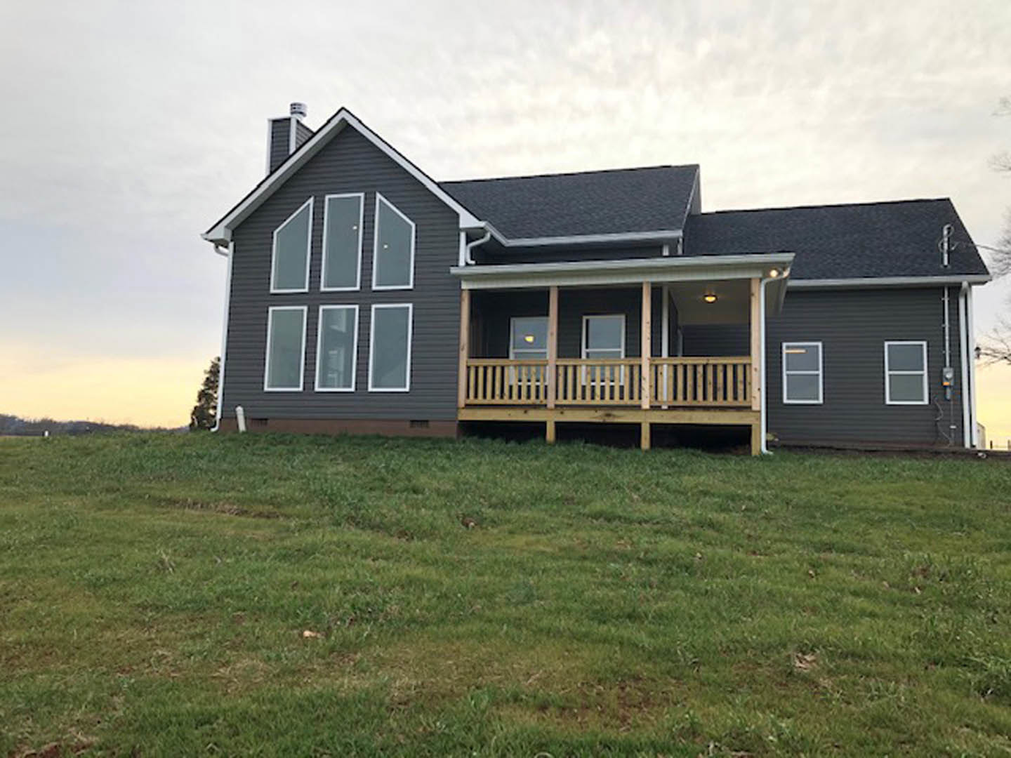 Two-story home with white siding and wooden porch, large windows with white frames, situated on a grassy hill bordered by a wooden fence under a partly cloudy sky