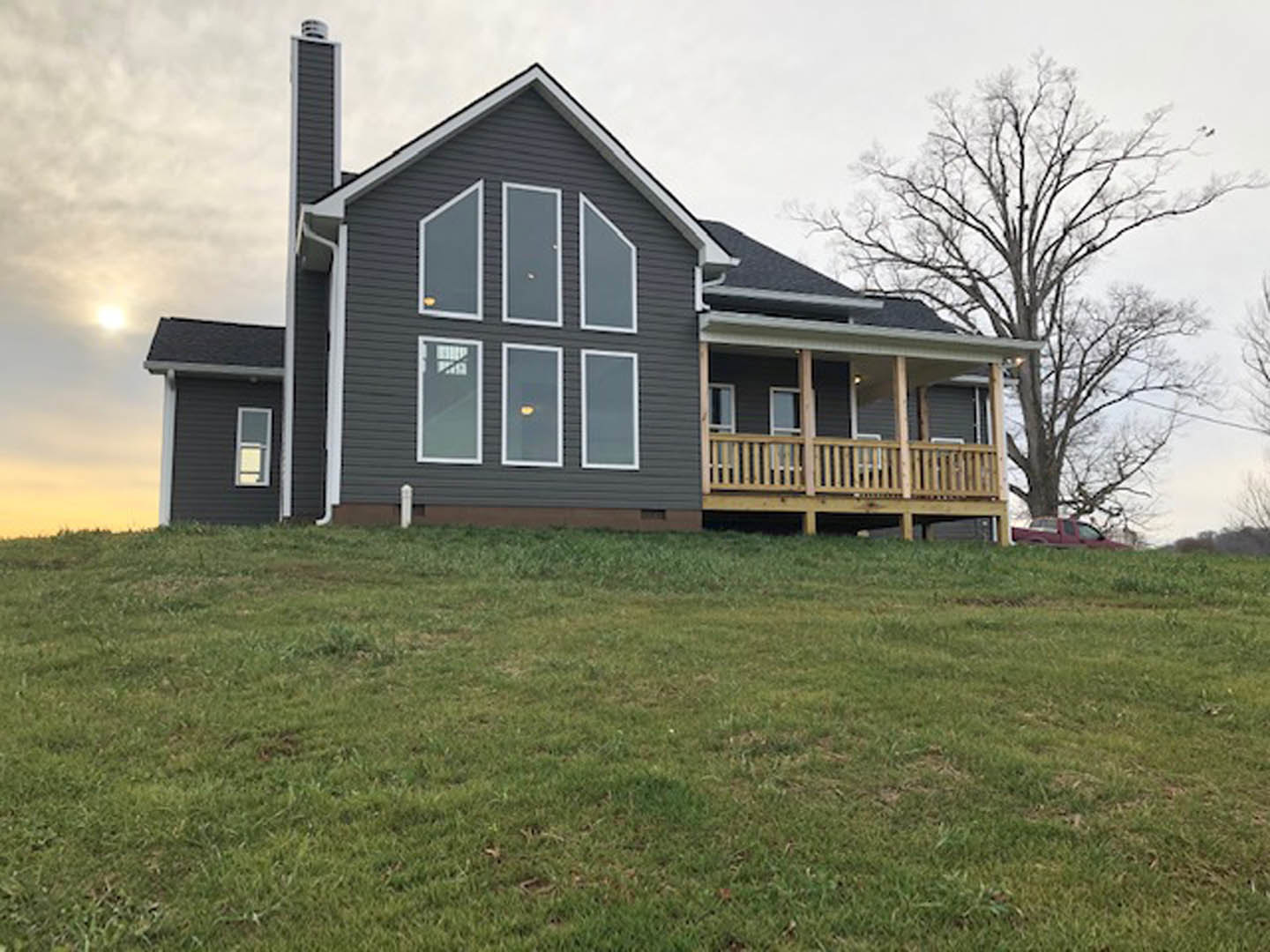 Modern two-story house with large windows and white trim, wooden deck with railing overlooking grassy hill, scattered trees and cloudy sky in background