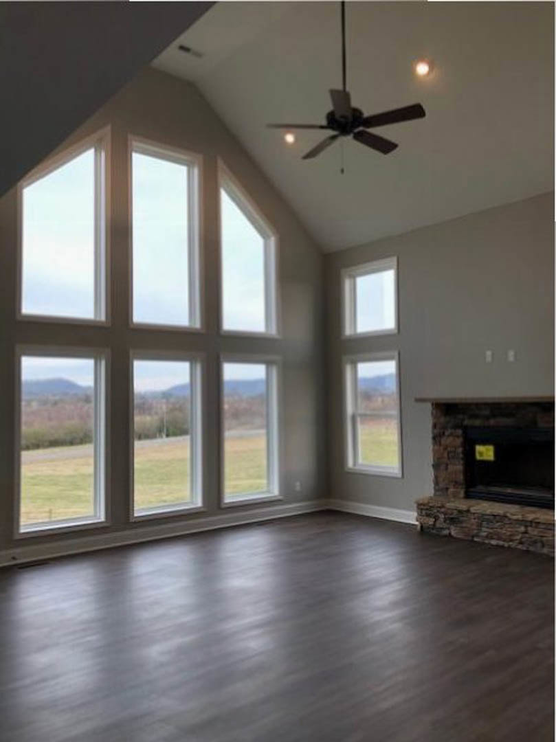 Living room with wood flooring, stone fireplace, ceiling fan with lights, and large window overlooking grassy field