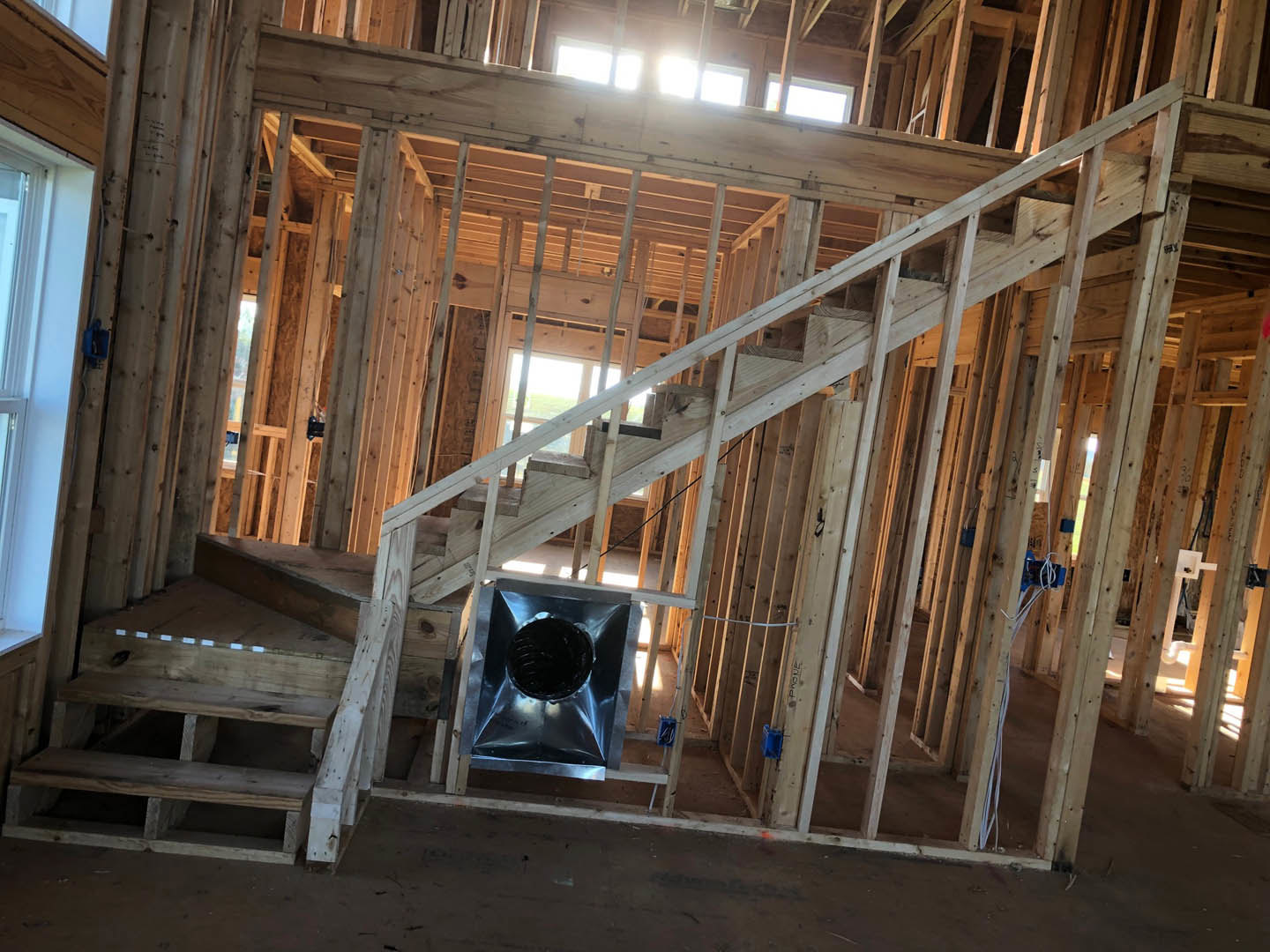 Exposed wood framing and beams inside a house under construction, with visible insulation, unfinished staircase, and construction materials scattered throughout.