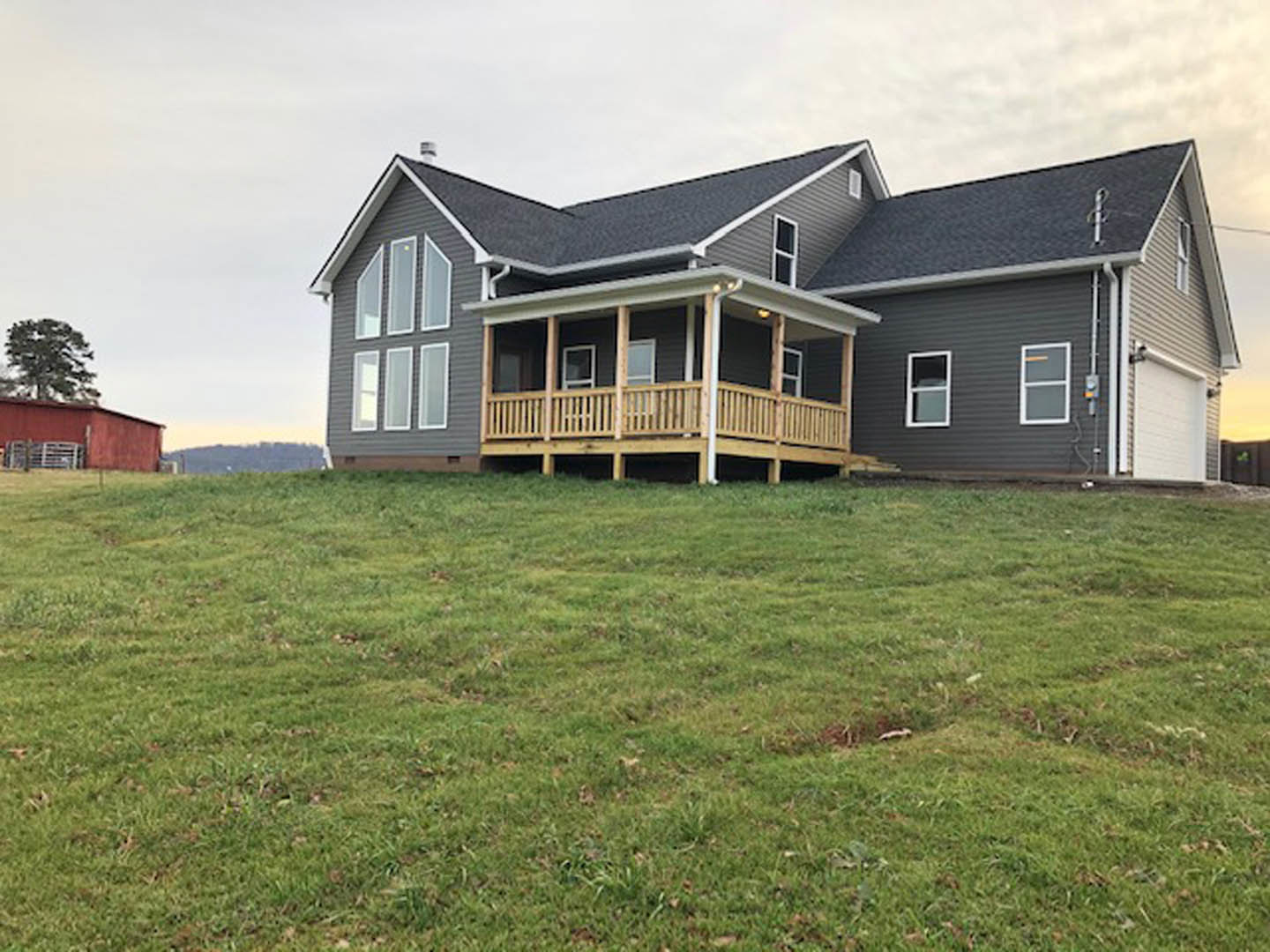 White siding house with side porch featuring wooden deck and railing, surrounded by green grass, windows, and cloudy sky in background