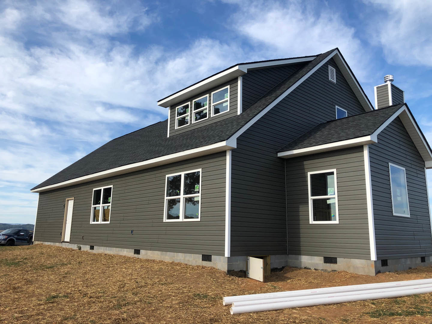 Modern cottage-style home with minimal windows, light-colored siding, white window frames, black truck parked on driveway, scattered white pipes on lawn, cloudy sky overhead
