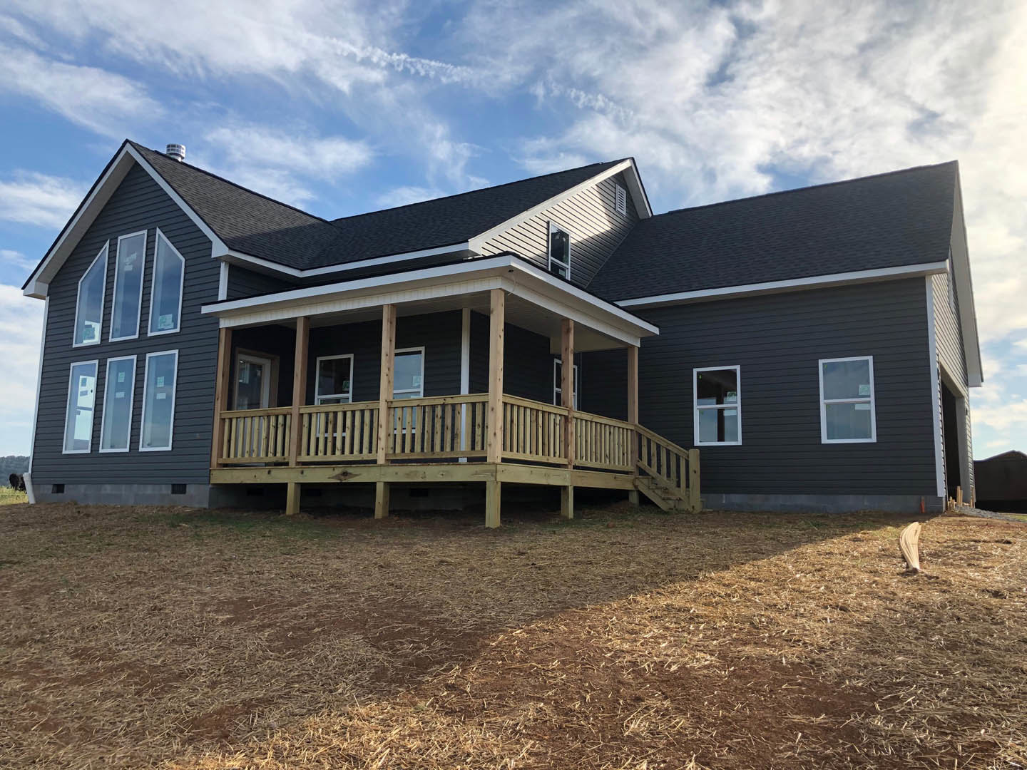 Two-story house with light-colored siding, covered front porch featuring white railings, wooden deck, white-framed windows, straw-covered yard, and partly cloudy blue sky