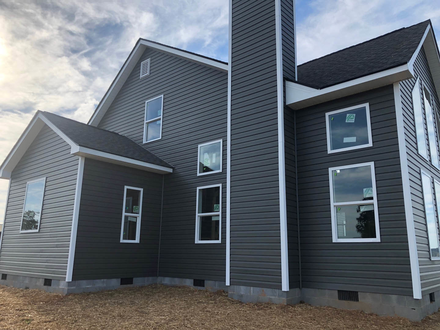 Gray siding house with white framed windows, green sticker on front window, brick chimney, tree reflected in glass, cloudy sky overhead