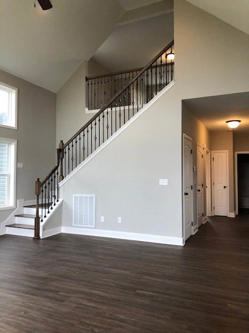 Dark wood staircase with black metal railings, white door with black handle, window with white shutters, sunlight streaming onto laminate flooring