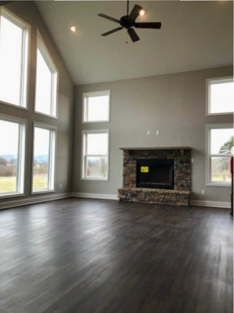 Living room with wood flooring, stone fireplace, ceiling fan with light, and large window; yellow sign on black surface near fireplace
