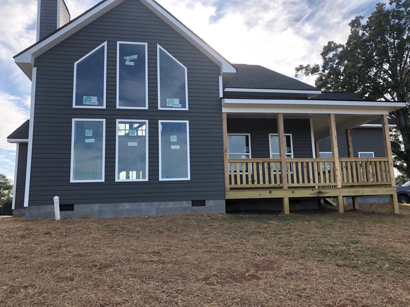 Two-story house with gray siding, multiple windows, covered front porch, wooden deck with railing, manicured lawn, and trees in the background