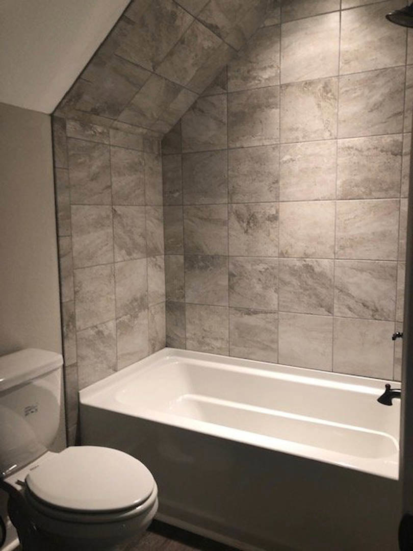 White bathroom with a built-in bathtub, two toilets side by side, light tile flooring, and neutral wall tiles