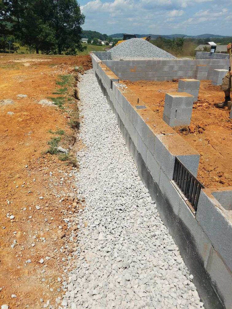 Concrete exterior wall with metal grate, grey block resting on soil, metal fence in foreground, tree and grass under blue sky with scattered clouds