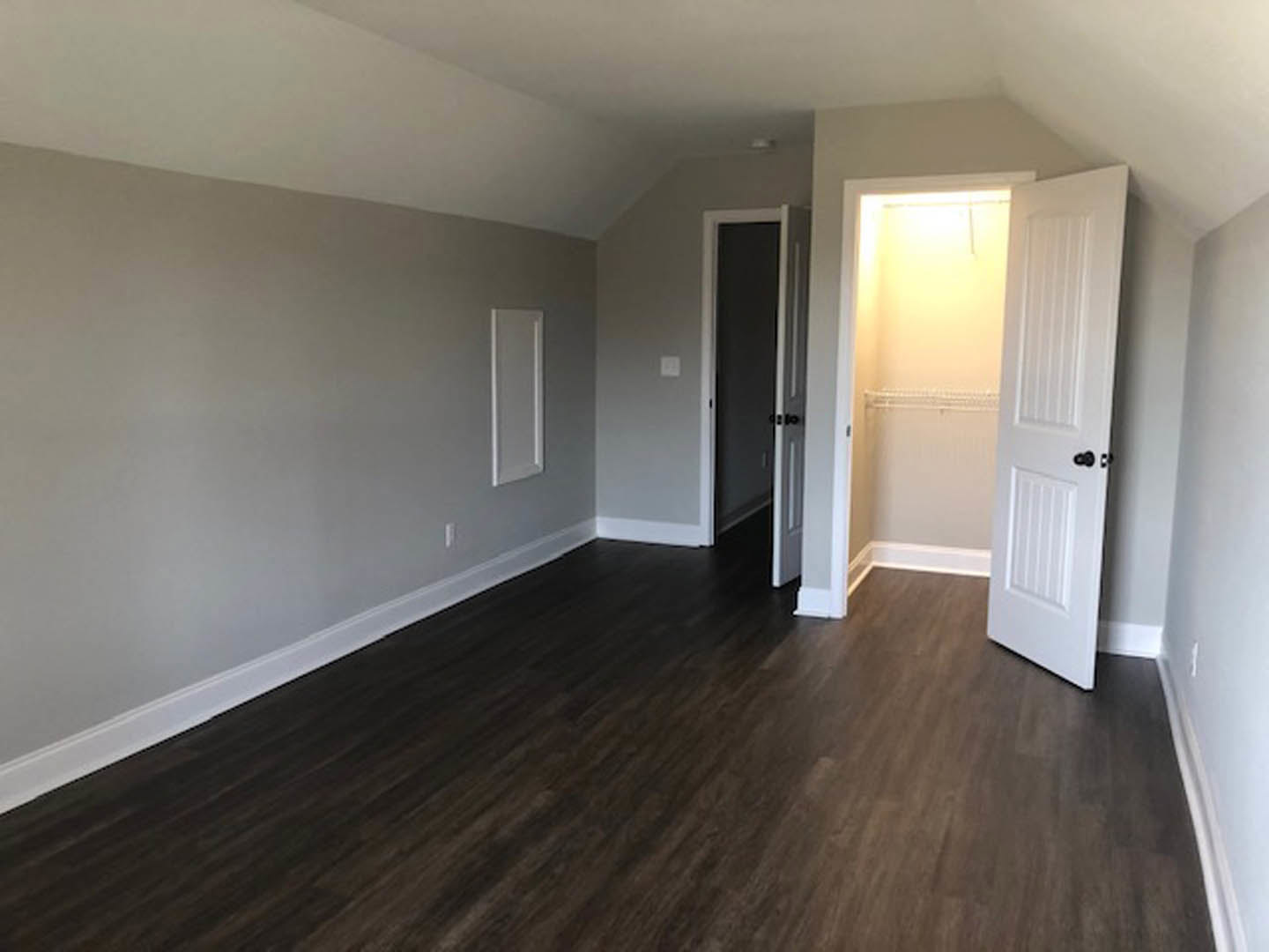 White paneled door with black hardware open to reveal a lit closet, dark wood laminate flooring, white walls, and trim in a residential interior.