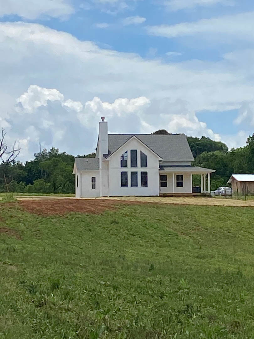 White farmhouse with chimney, surrounded by grassy hill and mature trees, set against a cloudy sky