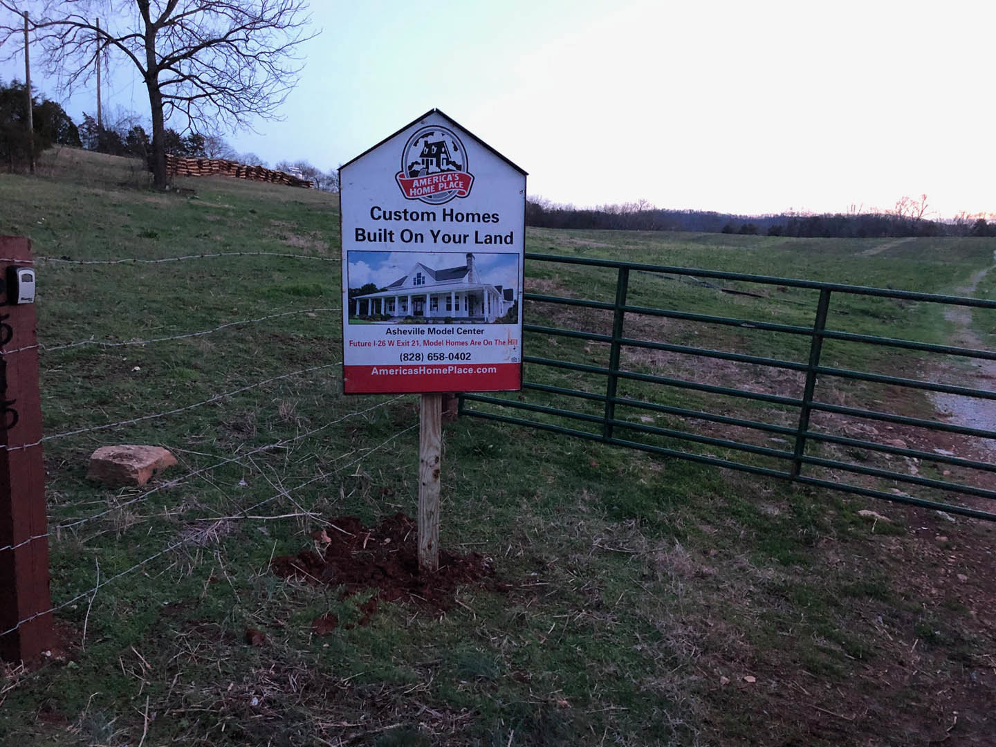 Barbed wire fence and leafless tree beside a large red rock in grassy field, sign with house illustration and text in foreground, blue sky overhead