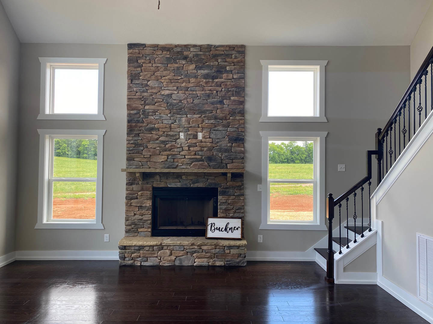 Stone fireplace with wooden mantel, black lettered sign above, large windows overlooking green field, hardwood floors, partial view of staircase railing