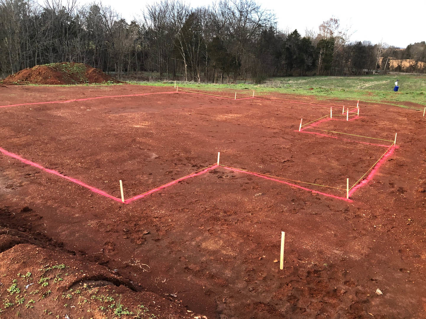 Dirt field with a red line marking the ground, grassy hill in the background, scattered trees under a pale sky