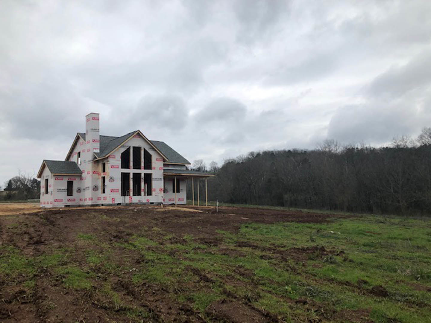 Wood-framed house under construction on a grassy dirt lot, surrounded by trees under a cloudy sky, red tape marking boundaries