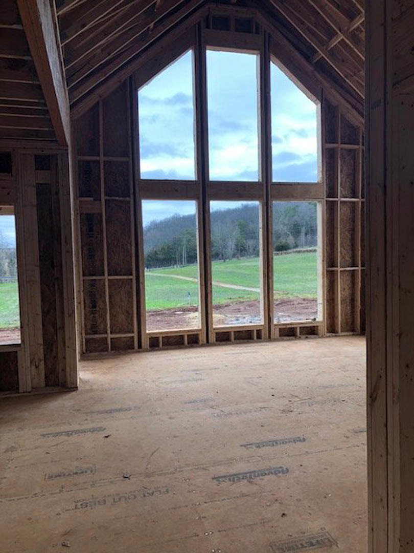 Spacious room featuring a large window overlooking a field and trees, wood flooring with black text, exposed wood paneling, and natural daylight illuminating interior finishes.