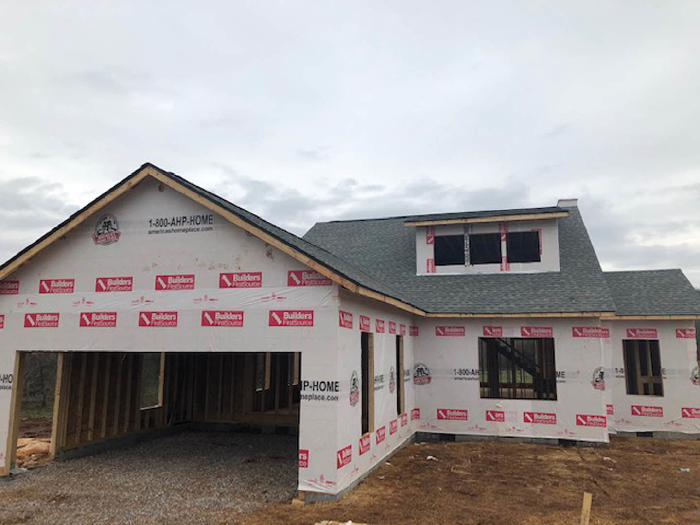 Framed house under construction with exposed plywood, red and white safety signs on exterior, unfinished siding, visible windows, cloudy sky overhead