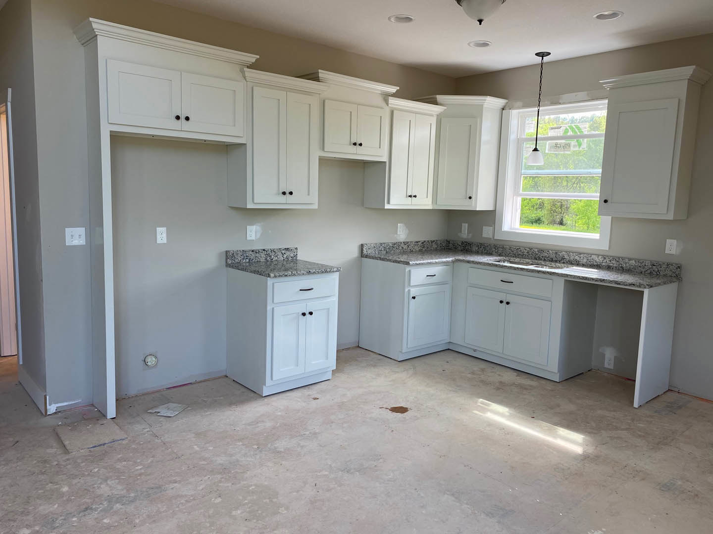 White kitchen cabinets with granite countertops, stainless steel appliances, window with natural light, stained floor, hanging ceiling chain, and white door in the background.