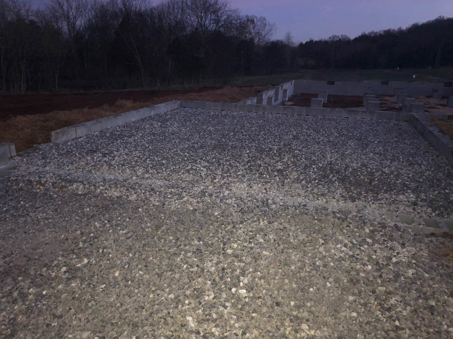 Concrete foundation surrounded by scattered rocks, gravel floor, and silhouetted trees against a dark nighttime forest sky