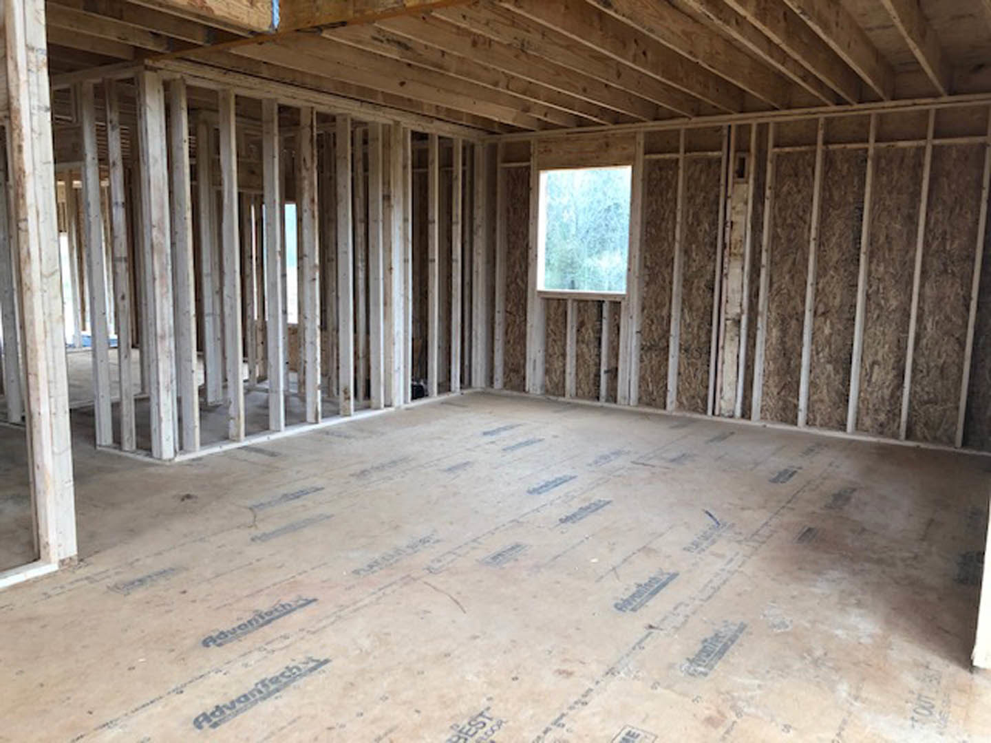 Room under construction with exposed wood framing, wooden ceiling beams, unfinished walls, and a window overlooking blurred trees.