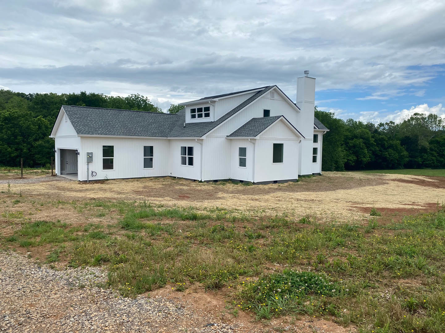 White farmhouse with black roof, expansive grassy yard, cloudy sky, and trees in the background