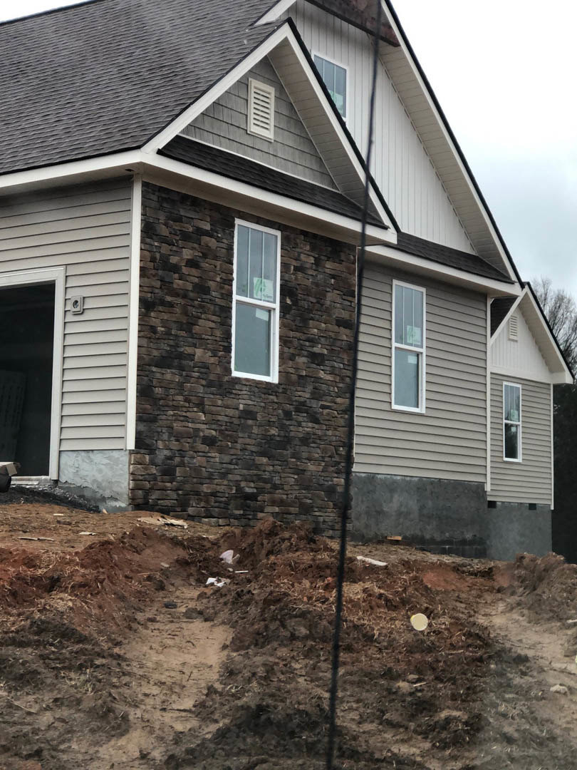 Two-story house with light siding, large windows, and a pile of dirt in the front yard; ladder leaning against exterior wall, construction materials scattered on bare ground.