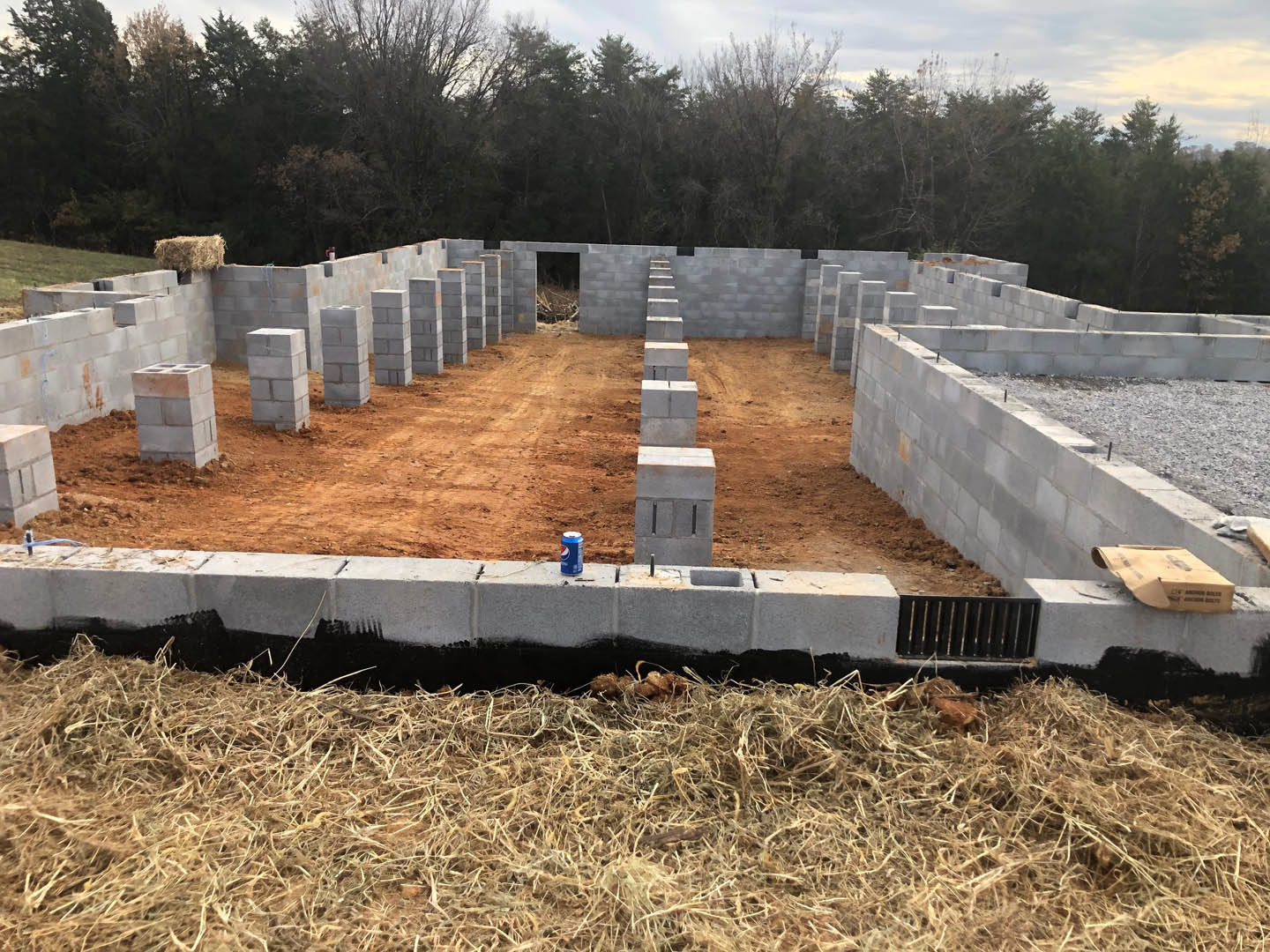 Concrete blocks and scattered hay on a residential building site, metal fence in foreground, grassy ground and trees in background under cloudy sky.